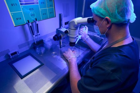Foto de Laborante en guantes estériles y mascarilla protectora mirando al microscopio, seleccionando óvulos sanos y preparándolos para la fertilización in vitro - Imagen libre de derechos