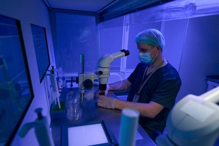 Foto de Hombre en uniforme estéril, mascarilla protectora, guantes y sombrero realizando investigaciones bajo microscopio en laboratorio con luz ultravioleta - Imagen libre de derechos