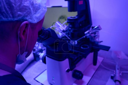 Foto de Trabajador de laboratorio del hombre concentrado mirando en lentes oculares del microscopio, llevando a cabo investigaciones, estudiando microcélulas en luz ultravioleta - Imagen libre de derechos