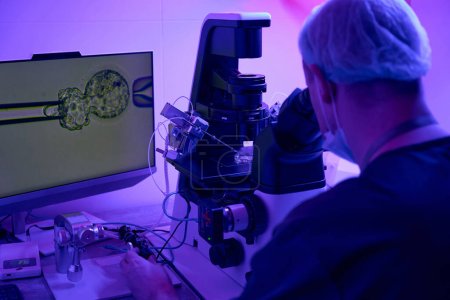 Embriólogo cultivando células en biolaboratorio utilizando micromanipulador, mirando sus acciones en pantalla digital conectada con microscopio