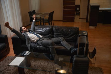 Téléchargez les photos : Un voyageur se repose de la route sur un canapé en cuir dans une chambre, à côté d'une valise de voyage - en image libre de droit