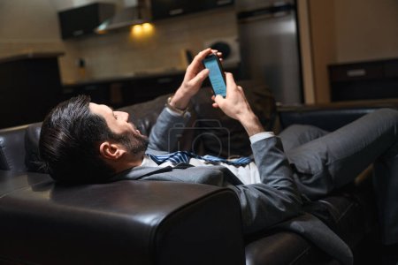 Foto de El huésped del hotel se está relajando en un sofá de cuero en la habitación, está usando un teléfono móvil - Imagen libre de derechos