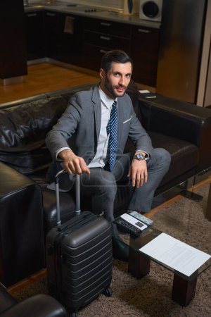 Foto de El huésped del hotel en un traje de negocios se sienta en un sofá de cuero, junto a una maleta de viaje - Imagen libre de derechos