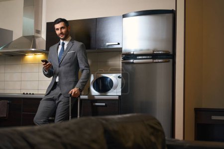 Foto de Hombre sonriente con teléfono móvil se encuentra en la zona de cocina de la habitación de hotel, junto a la maleta de viaje - Imagen libre de derechos