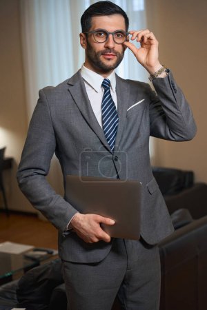Téléchargez les photos : Homme d'affaires se tient dans une chambre d'hôtel avec un ordinateur portable dans ses mains, un homme porte un costume élégant et des lunettes - en image libre de droit