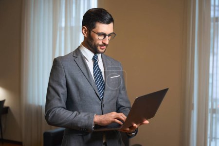 Téléchargez les photos : Élégante brune est debout dans une chambre d'hôtel avec un ordinateur portable dans ses mains, un homme porte un costume d'affaires élégant - en image libre de droit