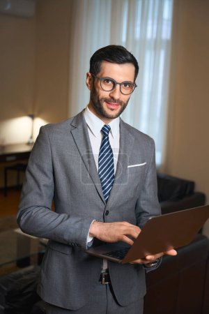 Téléchargez les photos : Belle brune en lunettes est debout dans une chambre d'hôtel avec un ordinateur portable dans ses mains, l'homme porte un costume d'affaires - en image libre de droit