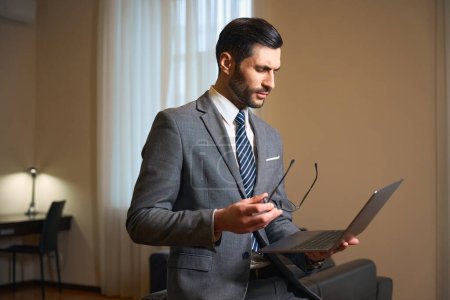Téléchargez les photos : Brune non rasée se tient dans une chambre d'hôtel avec un ordinateur portable et des lunettes dans ses mains, l'homme porte un costume d'affaires - en image libre de droit