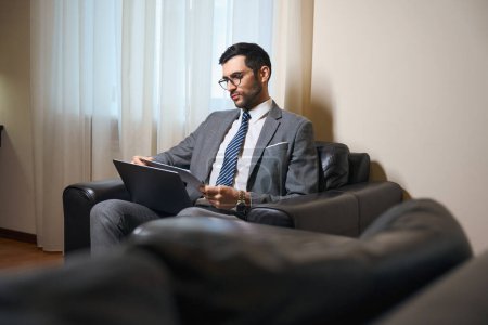 Téléchargez les photos : Homme en costume d'affaires et lunettes assis avec un ordinateur portable dans une zone de détente, il est assis dans une chaise en cuir - en image libre de droit