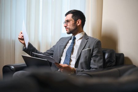 Photo for Man in a business suit and glasses is studying working documents in relax zone, he is sitting in leather chair - Royalty Free Image