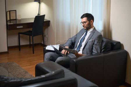 Téléchargez les photos : Homme d'affaires en costume d'affaires étudie les documents de travail dans une zone de détente, il est assis sur une chaise en cuir - en image libre de droit