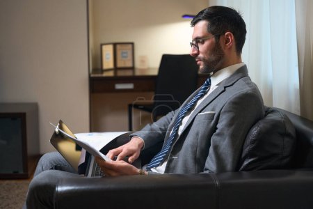 Téléchargez les photos : Beau homme en costume d'affaires étudie les documents de travail dans une zone de détente, il est assis dans une chaise confortable - en image libre de droit