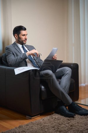 Téléchargez les photos : Homme d'âge moyen étudie les documents de travail dans la zone de détente, il est assis dans une chaise confortable - en image libre de droit