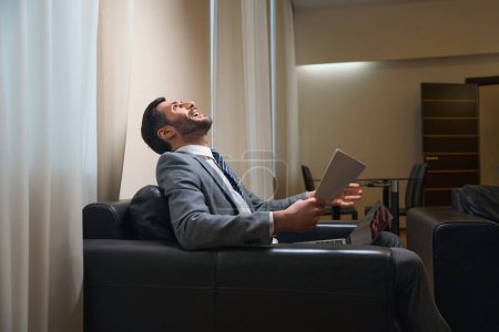 Foto de Hombre satisfecho en un traje de negocios se sienta en una silla de cuero, tiene documentos de trabajo y un ordenador portátil - Imagen libre de derechos