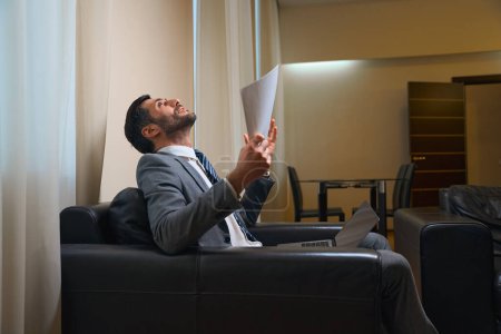 Téléchargez les photos : Homme heureux en costume d'affaires assis dans une chaise en cuir, il a des documents de travail et un ordinateur portable - en image libre de droit