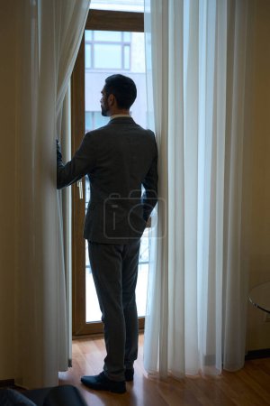 Téléchargez les photos : Un homme élégant en costume d'affaires se tient à la porte-fenêtre, le mâle admire la vue - en image libre de droit