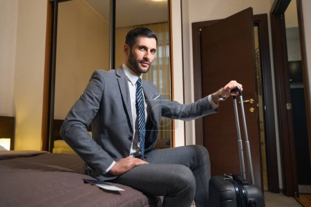 Photo for Traveling businessman with travel suitcase sits on a large bed in hotel room, he is dressed in a business suit - Royalty Free Image