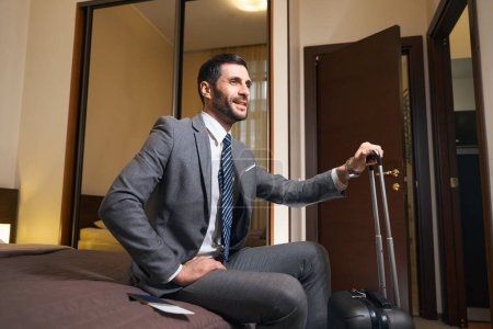 Foto de Hombre satisfecho con una maleta de viaje se sienta en una cama grande en una habitación de hotel, está vestido con un traje de negocios - Imagen libre de derechos