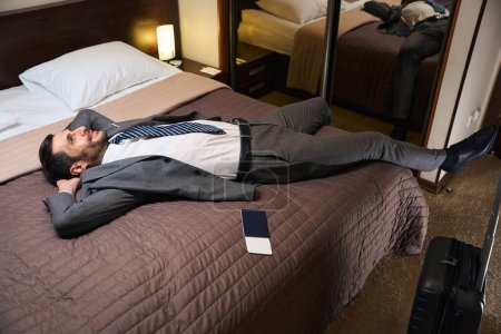 Foto de El hombre está descansando de la carretera en una cama grande en la habitación del hotel, junto a la maleta de viaje y un pasaporte - Imagen libre de derechos
