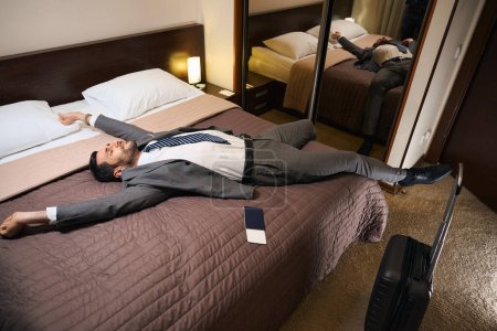 Foto de El huésped del hotel está descansando de la carretera en una cama grande en una habitación de hotel, junto a la maleta de viaje y el pasaporte - Imagen libre de derechos