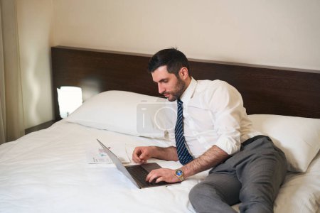 Photo for Unshaven middle-aged brunette reclining on the bed with working documents and a laptop - Royalty Free Image