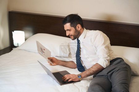 Photo for Unshaven brunette in business clothes reclining on the bed with working documents and a laptop - Royalty Free Image