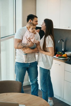 Photo for Loving dad holds small child in his arms and communicates with his wife, family is located in a bright kitchen - Royalty Free Image