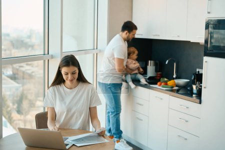 Téléchargez les photos : Père attentionné babysits bébé dans la cuisine pendant que maman travaille sur la paperasserie - en image libre de droit