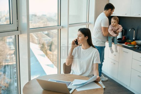 Photo for Beautiful female is talking on phone at the view window, next to her husband with a baby in her arms - Royalty Free Image