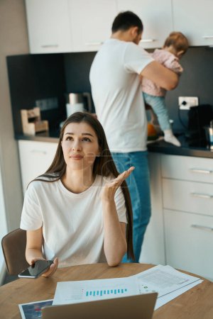 Photo for Young woman works remotely at home in the kitchen, next to her husband with a child in her arms - Royalty Free Image