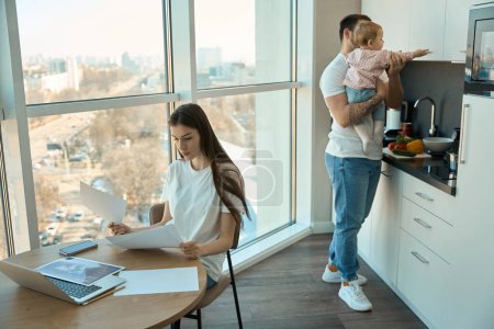 Téléchargez les photos : Jeune femme travaille à distance dans la cuisine à la fenêtre panoramique, à côté de son mari avec un enfant dans ses bras - en image libre de droit