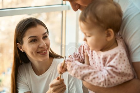 Téléchargez les photos : Mère moderne travaille confortablement à la maison, une petite fille dans les bras de son père joue avec un crayon - en image libre de droit