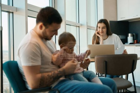 Téléchargez les photos : L'homme est babysitting un enfant dans la cuisine, à côté de lui est une mère heureuse avec ordinateur portable travaillant de la maison - en image libre de droit