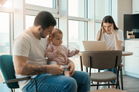Téléchargez les photos : Homme est babysitting un enfant dans la cuisine, à proximité est une mère fatiguée avec un ordinateur portable de travail de la maison - en image libre de droit