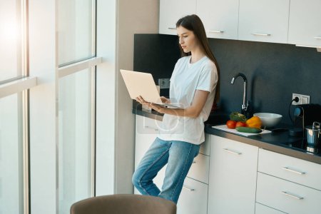 Photo for Young beautiful woman works from home on a laptop, the female stands at a large view window - Royalty Free Image