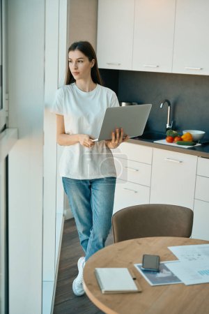 Photo for Charming long-haired woman stands, thinking, at a large view window, she works remotely on a laptop - Royalty Free Image