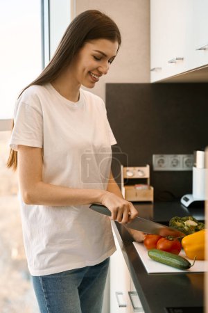 Téléchargez les photos : Agréable jeune femme coupe des légumes pour la salade dans une cuisine lumineuse, une femme se tient à la fenêtre - en image libre de droit