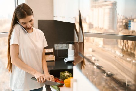 Photo for Nice young woman preparing vegetable salad and talking on the phone, she works remotely - Royalty Free Image
