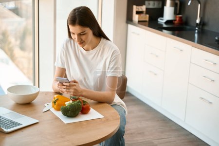 Foto de Morena hembra se sienta en la mesa de la cocina con un teléfono móvil en las manos, en la mesa un ordenador portátil y verduras - Imagen libre de derechos