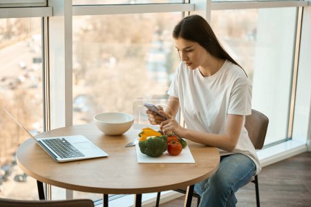 Téléchargez les photos : Belle femme est assise à la table de cuisine avec téléphone portable dans ses mains, sur la table est ordinateur portable et légumes - en image libre de droit