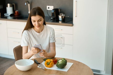 Téléchargez les photos : Jeune femme est assise à la table de la cuisine avec un téléphone portable dans ses mains, légumes sont sur la table - en image libre de droit