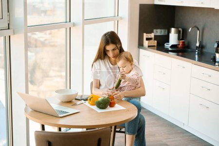 Téléchargez les photos : La mère attentionnée garde sa petite fille dans la cuisine, sur la table de cuisine il y a des ordinateurs portables, des légumes, des documents - en image libre de droit