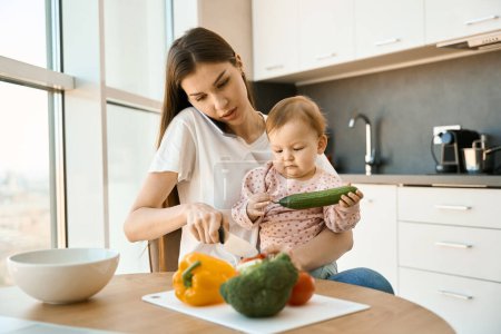 Téléchargez les photos : Jeune mère garde sa jolie petite fille dans la cuisine, la femme coupe des légumes - en image libre de droit