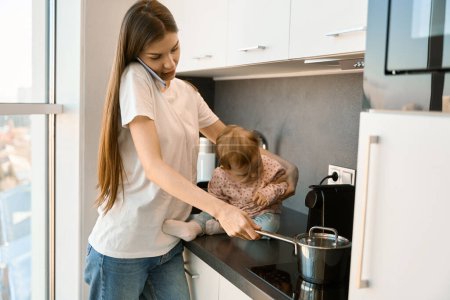 Photo for Young mother is preparing baby food and talking on the phone, her little helper is sitting on the kitchen cabinet - Royalty Free Image