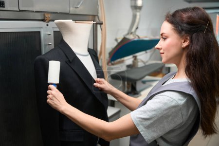 Téléchargez les photos : Femme qualifiée nettoyage à sec employé de bureau veste de nettoyage avec rouleau pelucheux collant, le portant au mannequin - en image libre de droit