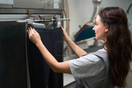 Trabajadora de servicio de lavandería mujer concentrada ajustando pantalones en maniquí de planchado, post-tratamiento después de la limpieza en seco