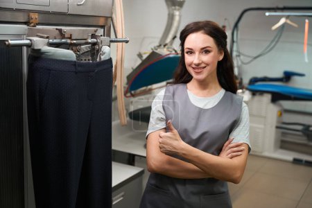 Photo for Happy wash house worker showing thumbs-up working with automatic ironing machine, processing clothes after cleaning - Royalty Free Image