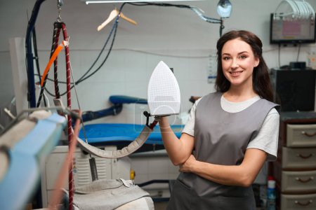 Foto de Mujer sonriente trabajadora de lavadero sosteniendo y mostrando utensilios eléctricos profesionales de planchado, servicio profesional de limpieza en seco - Imagen libre de derechos