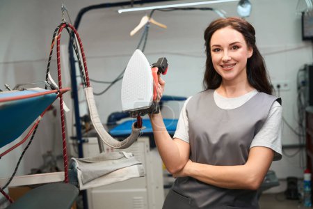 Téléchargez les photos : Femme opérateur de blanchisserie souriant tenant fer professionnel, travaillant dans un service de nettoyage à sec de haute qualité - en image libre de droit