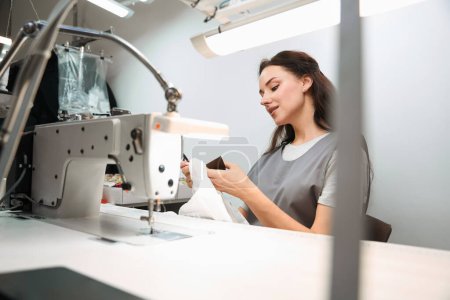 Foto de Mujer concentrada procesando ropa antes de la limpieza en seco, eliminando etiquetas sentadas en el lugar de trabajo con la máquina de coser - Imagen libre de derechos